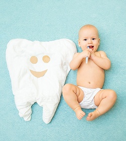 A baby with a toothbrush in its mouth and lying next to a pillow that looks like a tooth