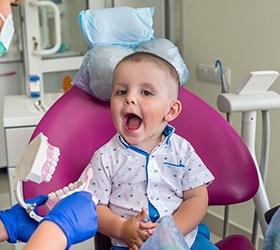 A baby sitting up in a dentist’s chair and opening his mouth as the dentist shows him how to brush his teeth